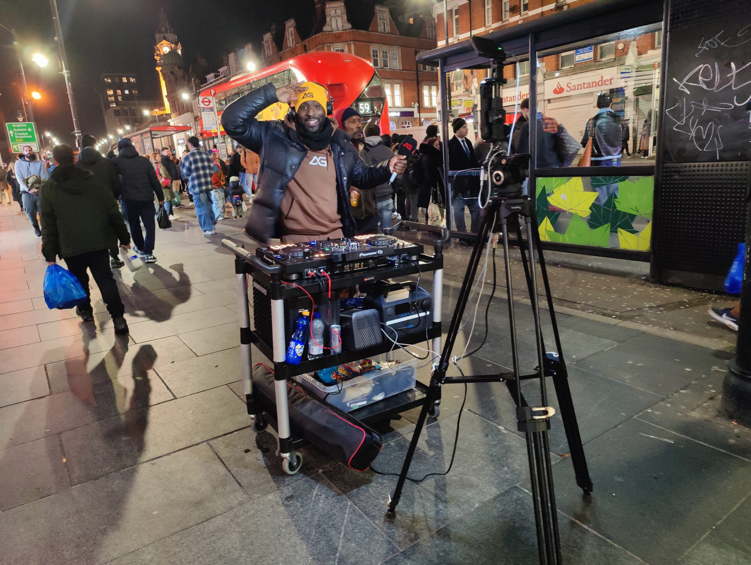 A man with black-skin wearing a heavy jacket and yellow bean hat. He is standing behind a trolley in the middle of the street in Brixton.

On the trolley is a complete DJ set-up - CD players, mixing desk, speakers.

In front of him is a tripod with a camera and lights.

He is saluting to the camera and smiling.