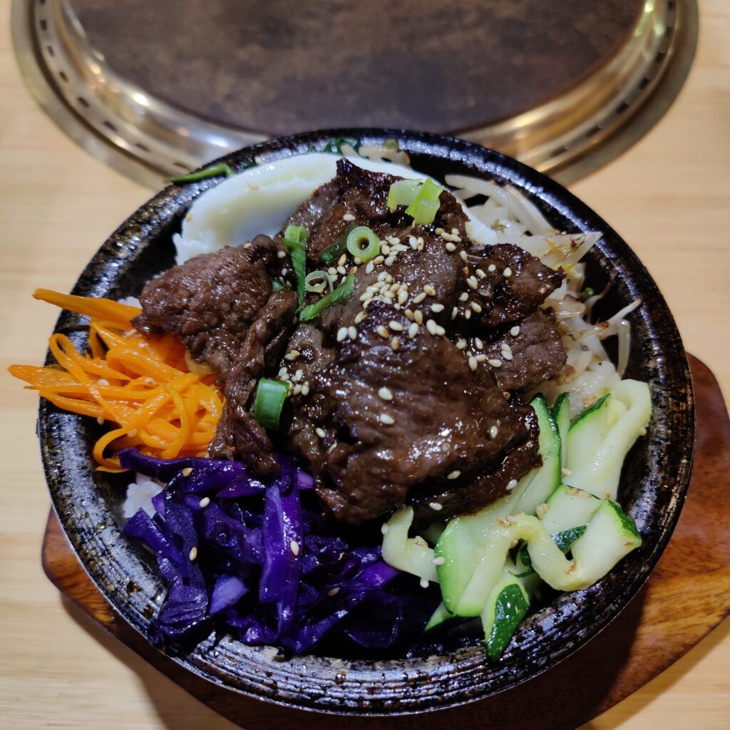 Dark coloured stone bowl containing colourful bibimbap - carrot, cucumber, rice, spring onion, beef bulgogi, cabbage, sprouts.